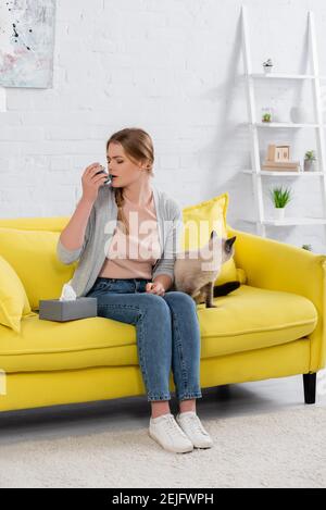 Young woman with allergy using inhaler near napkin and siamese cat Stock Photo