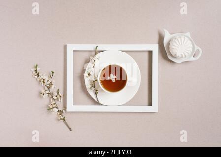 The concept of spring. A cup of black tea with apple blossoms in a white frame and marshmallows on a plate in the form of a teapot on a beige backgrou Stock Photo