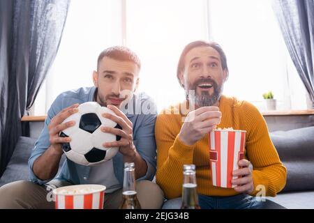 amazed man with popcorn watching football competition with hispanic son at home Stock Photo