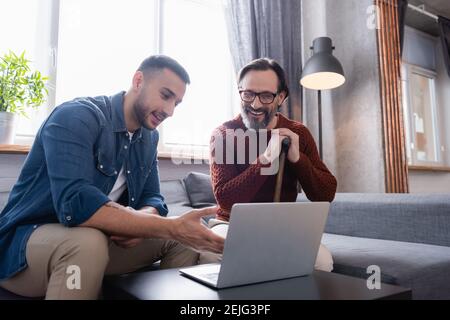 young hispanic man pointing at laptop near cheerful father sitting with walking stick Stock Photo