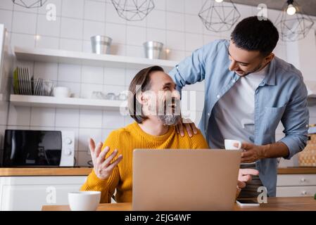 laughing man looking at young hispanic son while sitting near laptop in kitchen Stock Photo