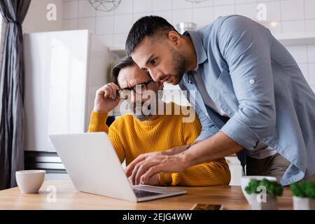 young hispanic man typing on laptop near thoughtful father adjusting eyeglasses, blurred foreground Stock Photo