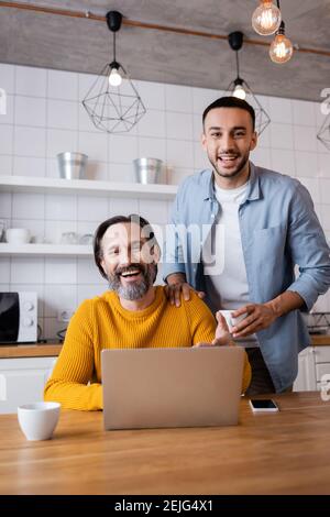 happy interracial father and son laughing at camera near laptop in kitchen Stock Photo