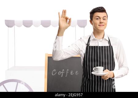 Young guy waiter holding an espresso coffee and gesturing ok sign in front of a coffee stand isolated on white background Stock Photo
