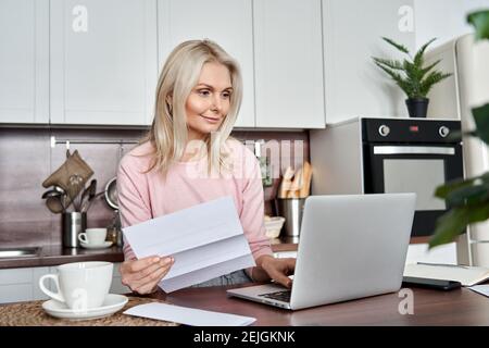 Middle aged mature woman holding paper bill or letter using laptop at home. Stock Photo