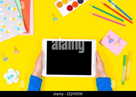 Online learning, back to school. Kid hands holding tablet. Headphones and school supplies on yellow background. Top view Stock Photo