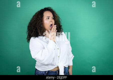 Young african american girl wearing white shirt over green background covering mouth with hand, shocked and afraid for mistake. surprised expression Stock Photo