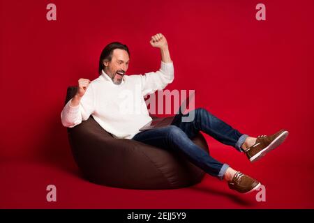Full size photo of mature excited crazy positive man raise fists in victory sit armchair isolated on red color background. Stock Photo