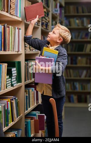 school boy taking books from shelves in library, with a stack of books in hands. child brain development, learn to read, cognitive skills concept Stock Photo