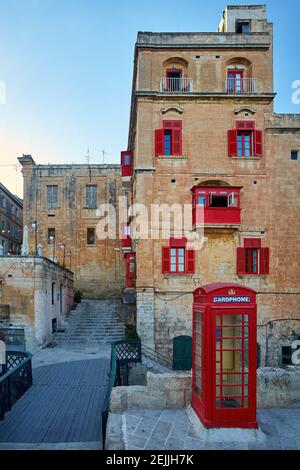 A deep red telephone kiosk on the street of the ancient city of Valletta in evening colors. Vertical, no people. Postcard. Holidays in Malta. Stock Photo