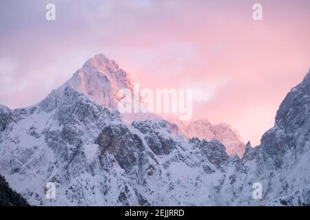 Beautiful close up shot of a pink glowing mountain tops in the Alps at sunset while wind is blowing snow off the mountain. Power of natural elements Stock Photo