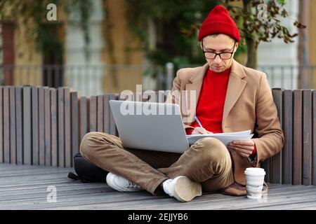 Serious hipster man reading documents, makes notes, remote work on laptop, sitting outdoors Stock Photo