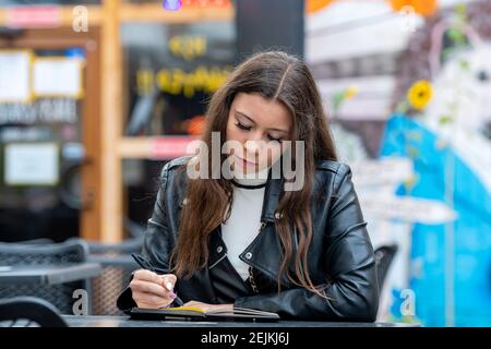 amazing optimistic young woman with long hair taking notes in notebook while sitting in outdoor cafe Stock Photo