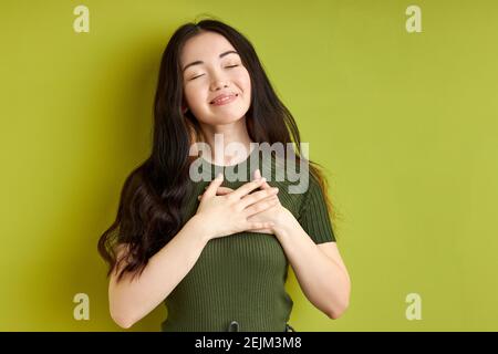 happy female expressing gratitude holding hands on chest with eyes closed, isolated over green studio background. portrait. people emotions concept Stock Photo