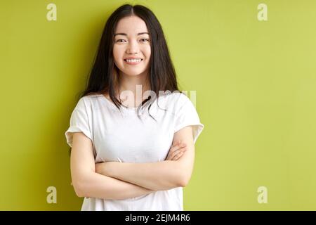 smiling woman with long black hair standing with arms folded, posing at camera isolated over green background, portrait Stock Photo