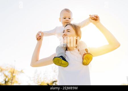 Happy young mother around her neck with her baby playing and having fun with her little son, on a sunny day Stock Photo