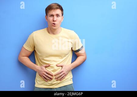 man feels stomachache isolated on the blue studio background, he holds hands on belly, stand feeling pain in stomach, wearing casual t-shirt. Stock Photo