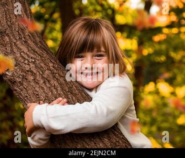 Little happy Girl hugging a tree with golden Autumn Foliage in the Background in a forest nature park on a sunny late summer day Stock Photo