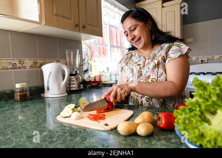 Happy Hispanic Mom Cooking Healthy Food - Mature Woman Cutting Fresh Vegetables In Her Kitchen - Woman Making Healthy Salad Stock Photo