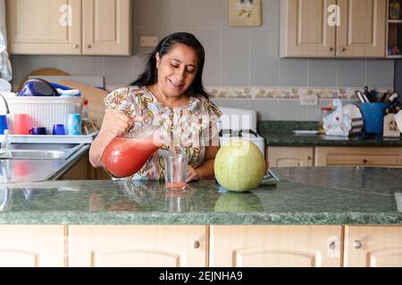 Happy hispanic mom pouring natural watermelon juice - Mature woman making natural watermelon soda -Woman pouring delicious fresh juice into glass on t Stock Photo