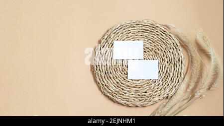Business cards white blank on the woven straw napkin, natural wheat spikelets on the beige neutral background. Flat lay, mockup  banner for displaying Stock Photo