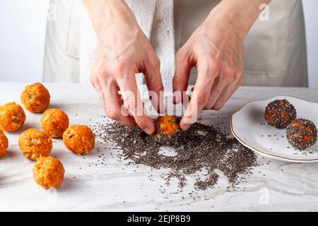 A woman chef is preparing carrot cake bliss bites in the kitchen. She dips the carrot balls into a pile of poppy and chia seeds and roll them to coat Stock Photo