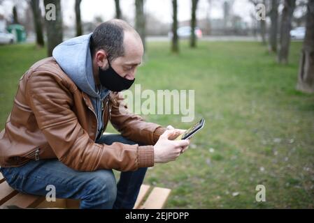 Caucasian man wearing a mask to protect himself from covid19 looks at his cell phone on a wooden park bench, open space. Stock Photo