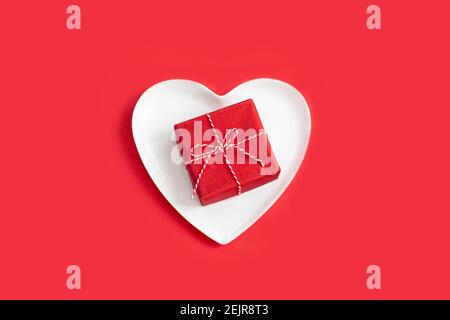 Small red gift box on a white heart-shaped plate. holidays table setting. Valentines day gift Stock Photo
