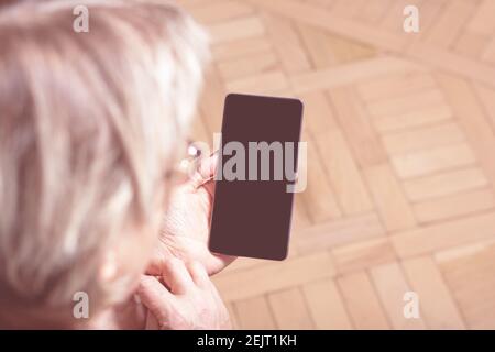 Elderly womans hand holding mobile cell phone. Over shoulder. Old woman looking at cell phone. Close-up. Blanck mobile phone screen. Display media.  Stock Photo