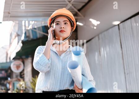 architect woman use mobile phone call with enginner or project manager between survey and checking at site construction. Architect woman hold blueprin Stock Photo