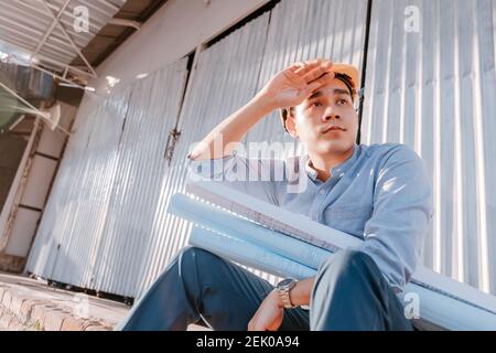 Architecture man wipe the sweat and him sitting on the ground and hold blueprints design plan and wearing safety helmet at site construction in hot we Stock Photo