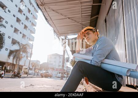 Architecture man wipe the sweat and him sitting on the ground and hold blueprints design plan and wearing safety helmet at site construction in hot we Stock Photo