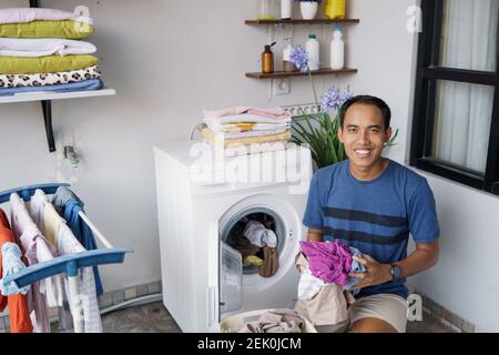 handsome asian househusband putting dirty clothes one by one into washing machine and doing laundry at home Stock Photo