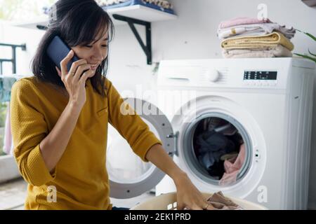 woman in front of the washing machine doing some laundry loading clothes inside while on phone call Stock Photo