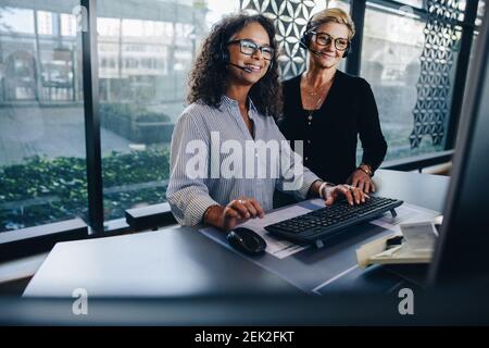 Smiling call center workers at the office desk. Two female coworkers wearing headset working together looking at computer monitor. Stock Photo
