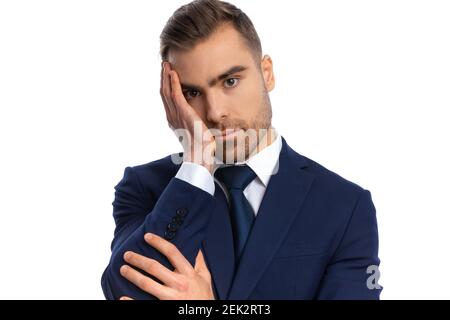 unhappy young guy in navy blue suit holding hand to face, having a headache and posing isolated on white background in studio Stock Photo