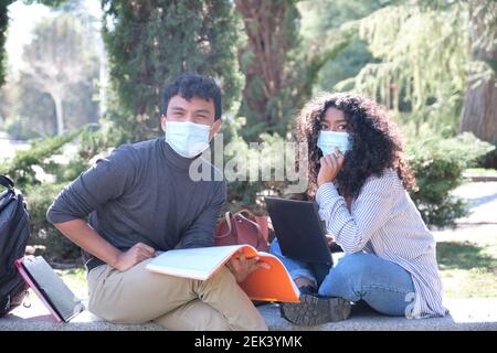 Two latin students wearing protective face mask studying together sitting on a bench outdoor. New normal at university campus. Stock Photo