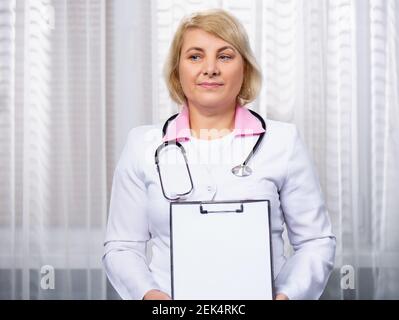 Senior female healthcare worker looking into distance, showing white blank clipboard for your personal message, advice or medical advertisement Stock Photo