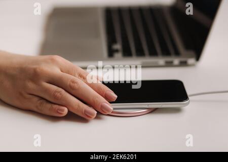Female hand tup phone on wireless charging. Work place near at the laptop. Top view Stock Photo
