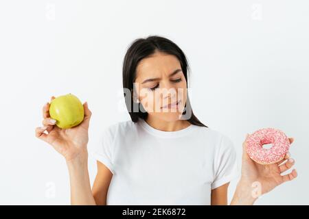 Young woman doubts what to choose healthy food or sweets holding green apple and donut in hands standing on white studio background. Stock Photo