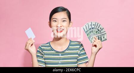 Portrait of asian beautiful woman 20s holding fan of money dollar banknotes and credit card isolated over pink background Stock Photo