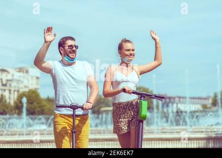 Happy couple with open face mask on vacation having fun driving electric scooter in city centre while greeting friends Stock Photo