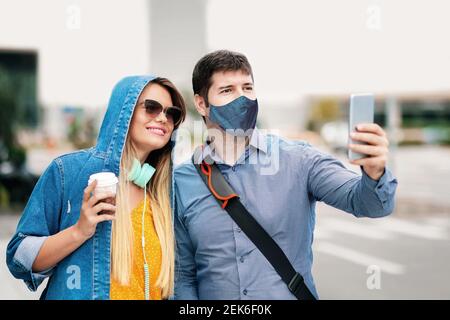 Happy boyfriend and girlfriend having fun taking selfie with face mask on city street Stock Photo