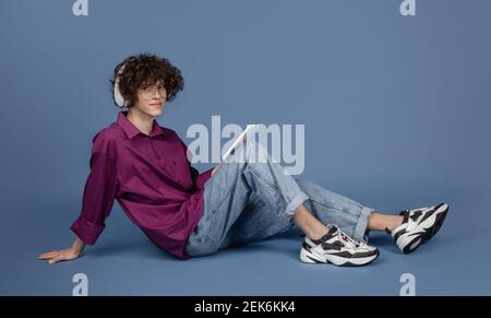 Listening to music. Caucasian young handsome man's portrait on blue studio background with copyspace. Male curly model with devices. Concept of human emotions, facial expression, sales, ad, fashion. Stock Photo
