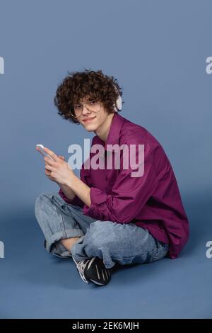 Listening to music. Caucasian young handsome man's portrait on blue studio background with copyspace. Male curly model with devices. Concept of human emotions, facial expression, sales, ad, fashion. Stock Photo