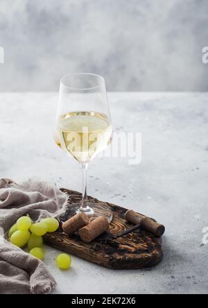 Glass of white homemade wine with corks, corkscrew and grapes on wooden board with linen cloth on light table background. Stock Photo