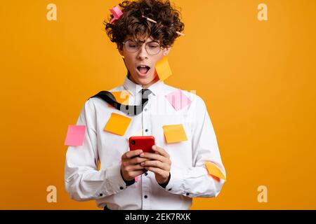 Shocked frustrated young man covered in post it stickers wearing white shirt using mobile phone isolated over yellow background Stock Photo