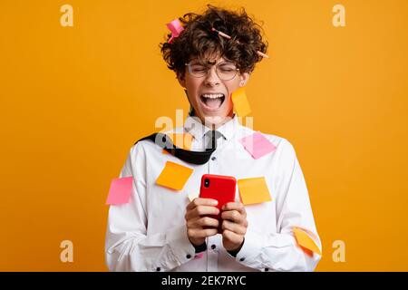Shocked frustrated young man covered in post it stickers wearing white shirt using mobile phone isolated over yellow background Stock Photo