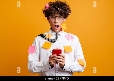 Shocked frustrated young man covered in post it stickers wearing white shirt using mobile phone isolated over yellow background Stock Photo