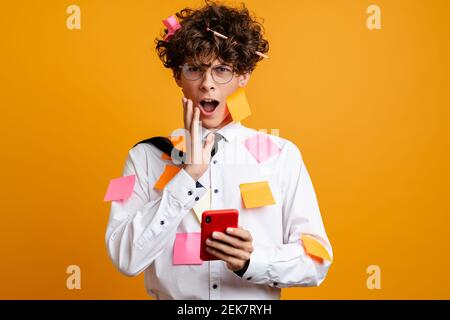 Shocked frustrated young man covered in post it stickers wearing white shirt using mobile phone isolated over yellow background Stock Photo